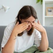 A person sitting on a couch rubs their eyes with both hands, appearing tired or stressed.