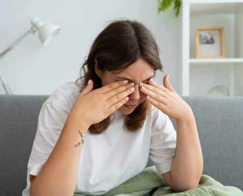 A person sitting on a couch rubs their eyes with both hands, appearing tired or stressed.