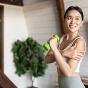 Woman in workout clothes holding a green dumbbell and touching her shoulder, standing indoors in a bright room with a plant in the background.