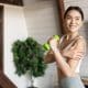 Woman in workout clothes holding a green dumbbell and touching her shoulder, standing indoors in a bright room with a plant in the background.