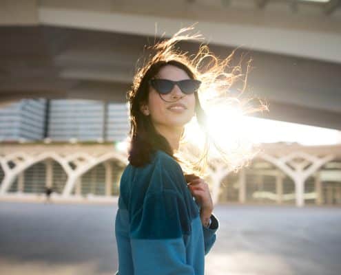 Woman wearing sunglasses and a blue jacket stands outdoors under a bridge, with sunlight shining behind her and her hair blowing in the wind.