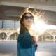 Woman wearing sunglasses and a blue jacket stands outdoors under a bridge, with sunlight shining behind her and her hair blowing in the wind.