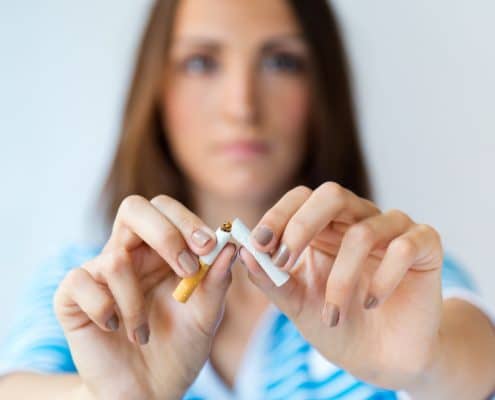 A woman in a blue-striped shirt breaks a cigarette in half with both hands, with her face out of focus in the background.