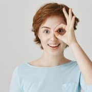 Young person with short brown hair and freckles smiles, making a circle with their fingers and holding it over one eye, standing against a plain light background.