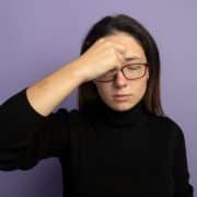 A person wearing glasses and a black sweater stands against a purple background, eyes closed, with one hand pressed to their forehead.
