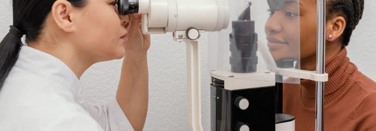 An eye care professional examines a patients eye using a slit lamp during an eye check-up. Both are separated by a clear plastic divider.