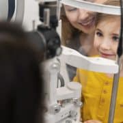 A young boy in a yellow shirt sits at an eye exam machine with a smiling adult behind him and an optometrist in front.