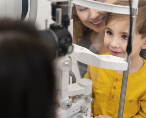 A young boy in a yellow shirt sits at an eye exam machine with a smiling adult behind him and an optometrist in front.