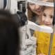 A young boy in a yellow shirt sits at an eye exam machine with a smiling adult behind him and an optometrist in front.