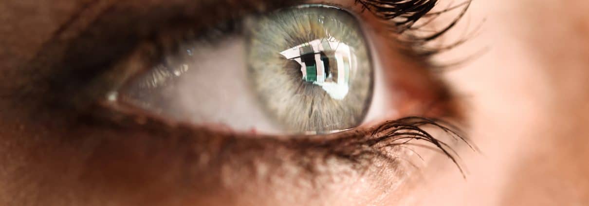 Close-up of a human eye with long eyelashes, hazel-green iris, and a reflection of a window in the pupil.