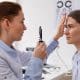 An eye doctor examines a woman’s eyes using an ophthalmoscope in a clinic, with an eye chart visible in the background.